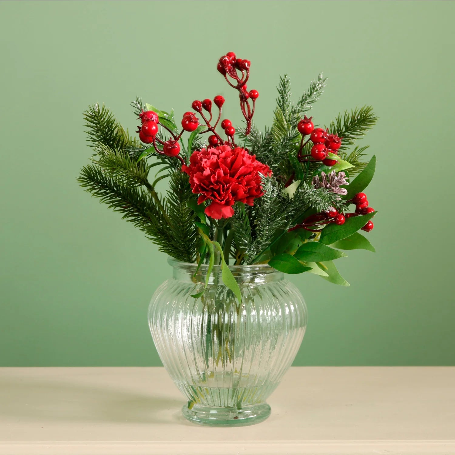 A festive artificial floral arrangement featuring a bright red carnation, red berries, green pine branches, and green leaves, presented in a ribbed clear glass vase on a light beige surface against a sage green background.