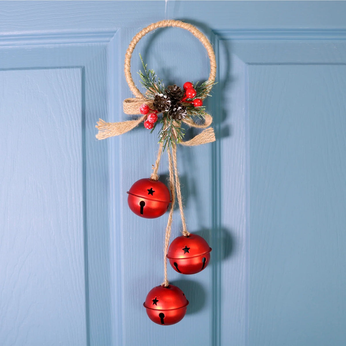 A rustic Christmas wreath hanging on a light blue paneled door. The wreath is made of natural jute rope and decorated with red berries, a small pinecone, green pine needles, and three hanging red bells.