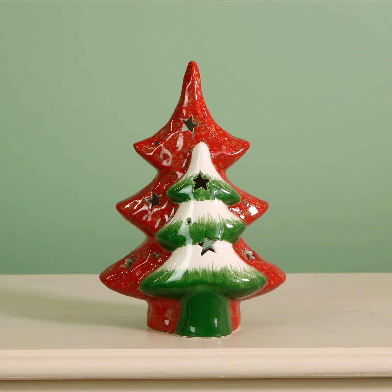 A ceramic Christmas tree-shaped candle holder with a red outer tree, a green tree with white snow-like accents, and star-shaped cutouts, displayed on a white surface against a pale green background.