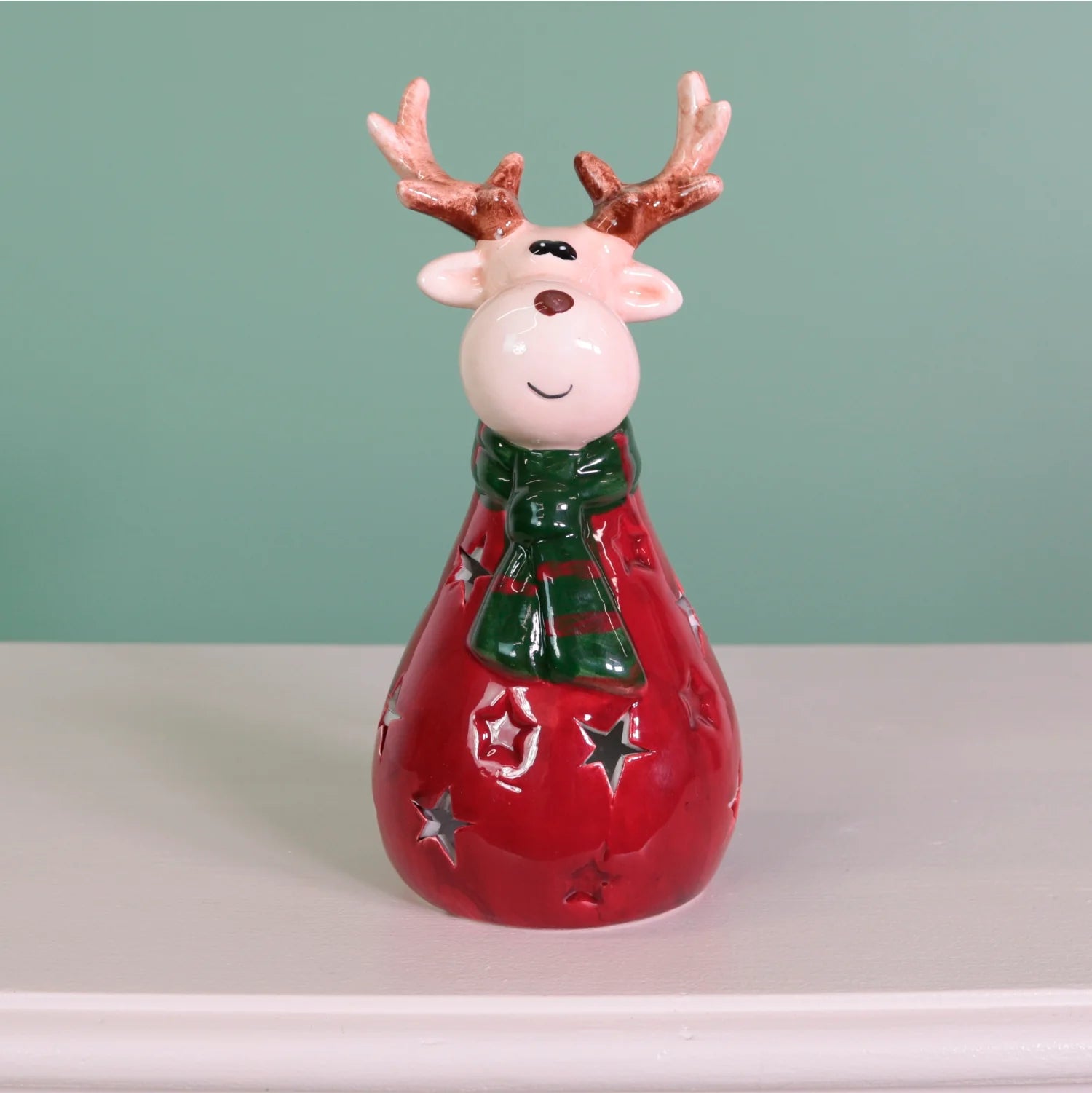 A ceramic Christmas reindeer figurine with brown antlers, a green and red striped scarf, and a red body decorated with silver star cutouts, standing on a white surface against a mint green background.
