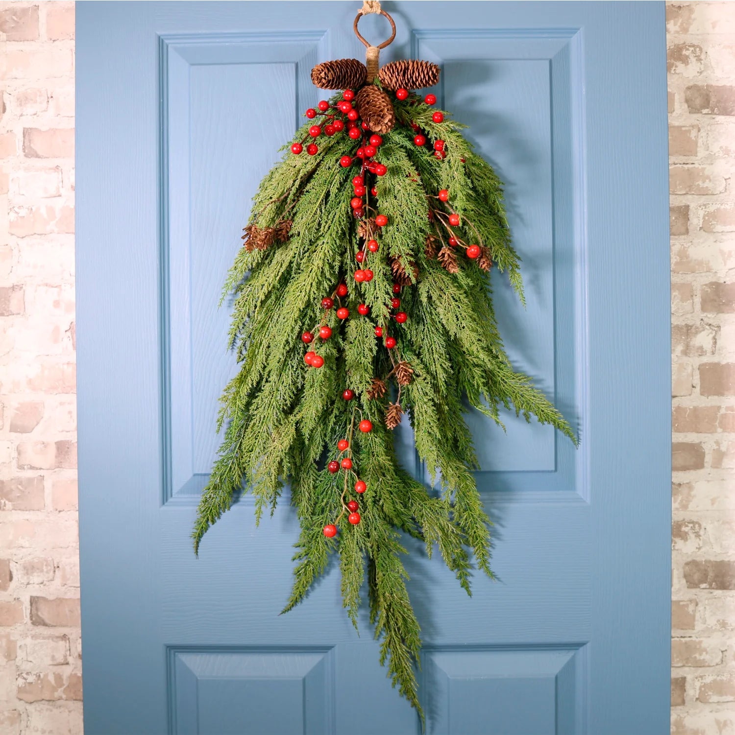 A hanging holiday wreath made of green evergreen branches, red berries, and brown pinecones, displayed on a blue front door with a brick wall visible on both sides.