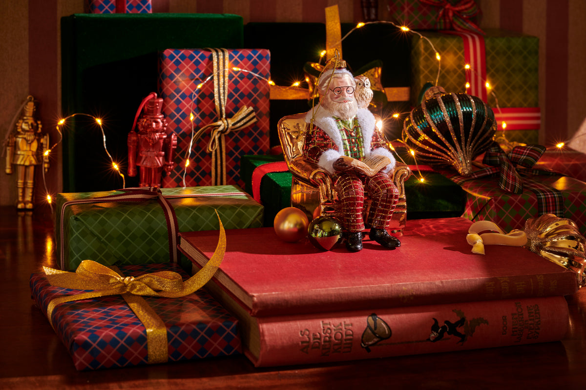 A festive holiday scene with a Santa Claus figurine seated on a red book, surrounded by wrapped Christmas presents, ornaments, string lights, and decorative figures on a wooden surface.