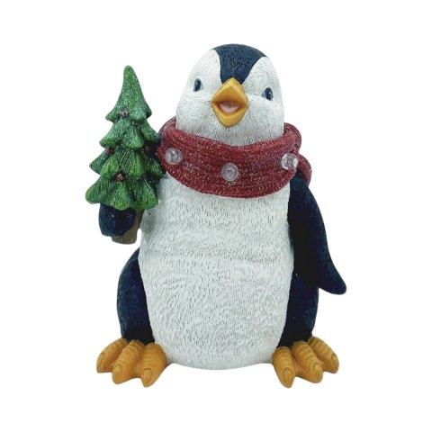 A penguin figurine with a white and black body, yellow beak and feet, wearing a red glittery scarf with silver ornaments and holding a small green Christmas tree, isolated on a white background.