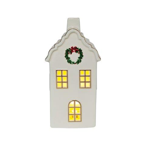 A white ceramic Christmas house ornament with glowing yellow windows and a green wreath with a red bow on the roof, isolated on a white background.