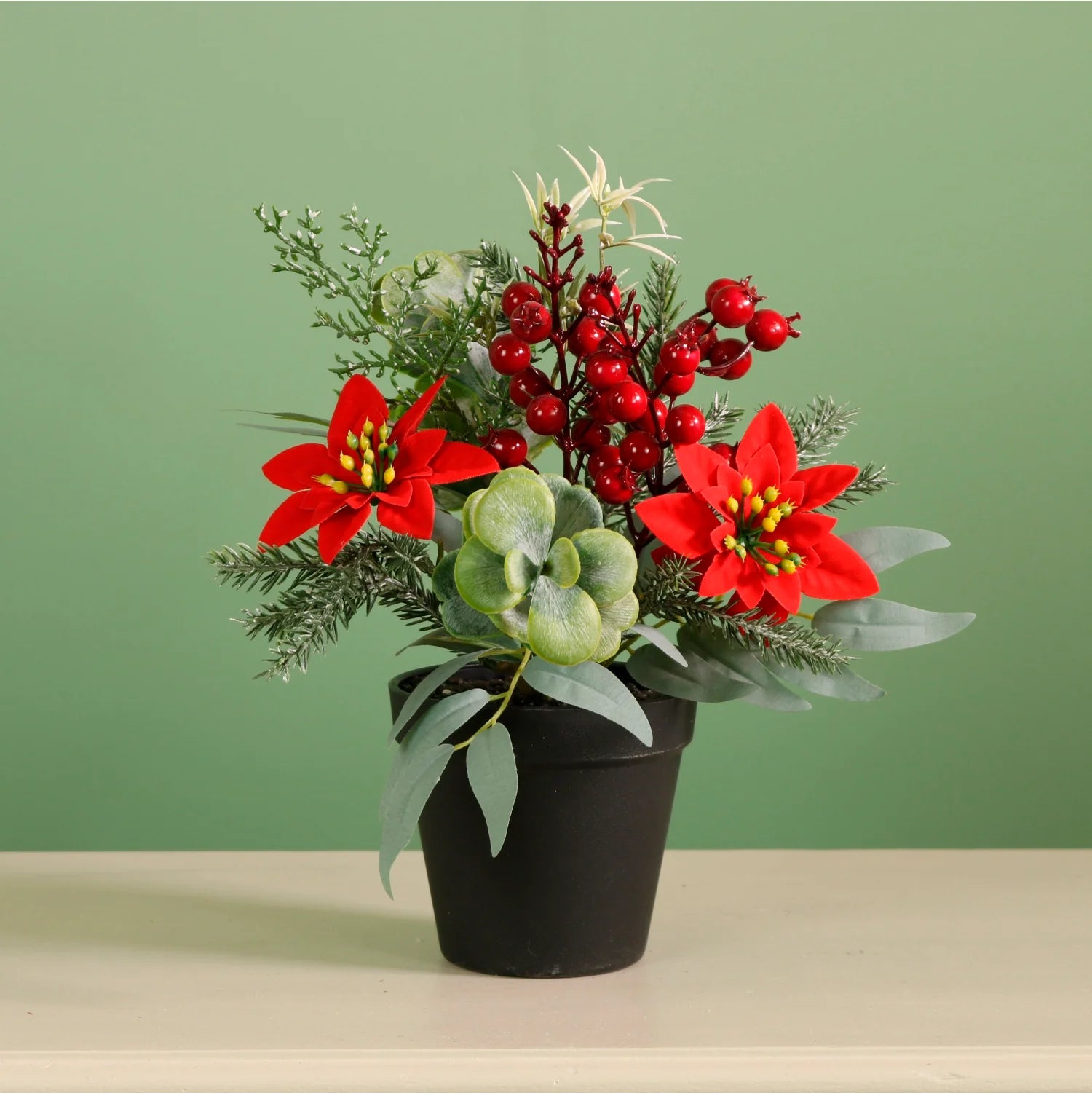 A festive artificial holiday floral arrangement in a small black pot, featuring red poinsettias, red berries, green foliage, and eucalyptus leaves, set against a solid sage green background.