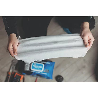A person holding up a large, light gray rectangular fabric or material sample, with a blue packaged product and tools visible in the blurred background on a light wood floor.