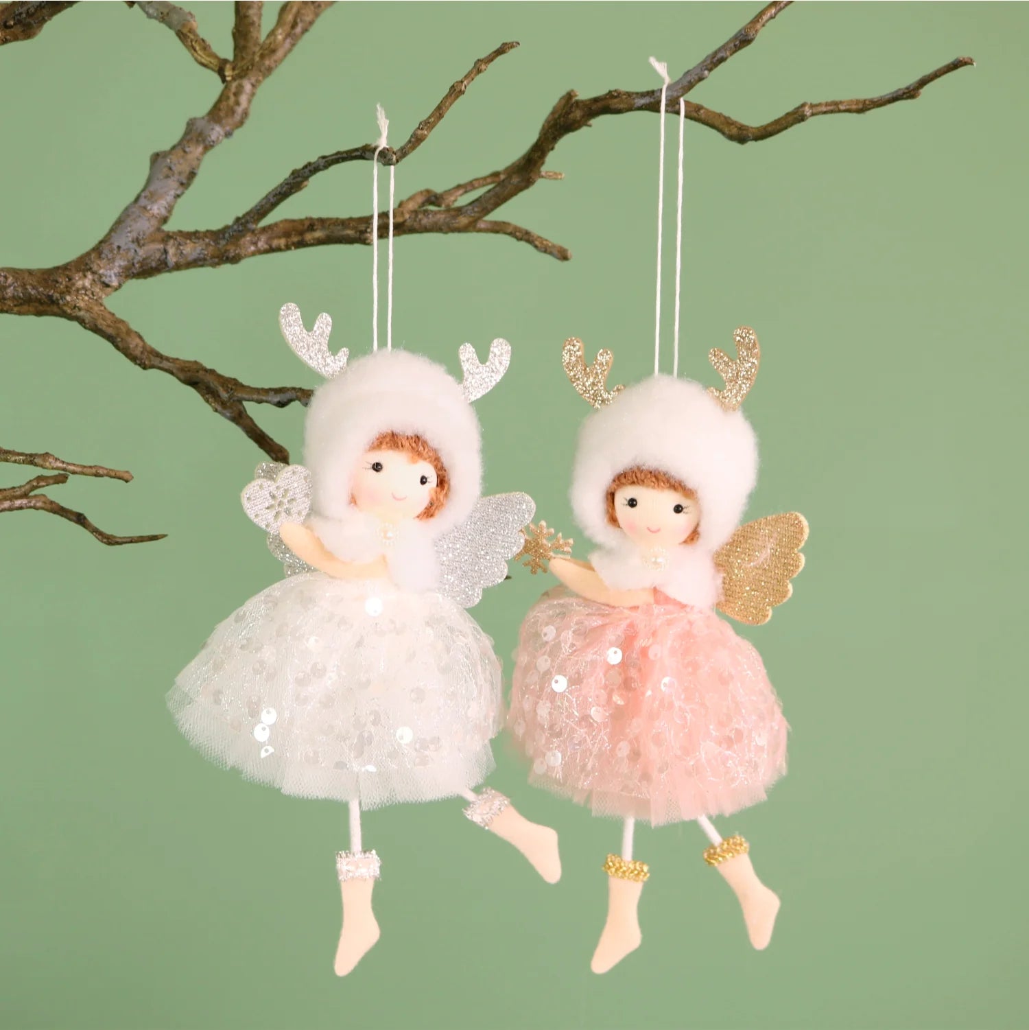Two hanging angel ornaments with fluffy white and pink dresses, glittery wings, and antler hats, suspended from a tree branch against a pale green background.