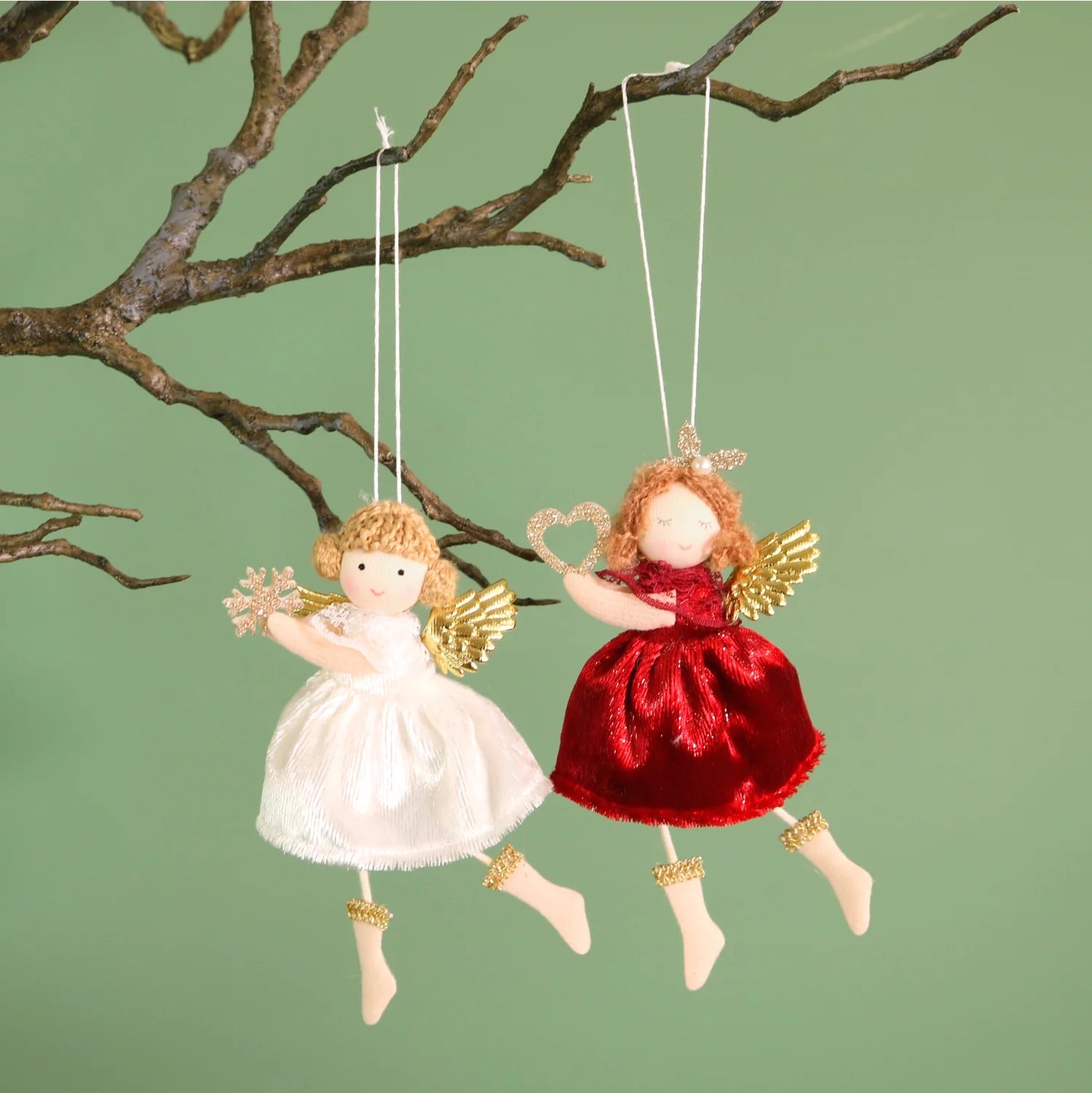 Two angel ornaments, one white and one red, hanging from branches against a green background.