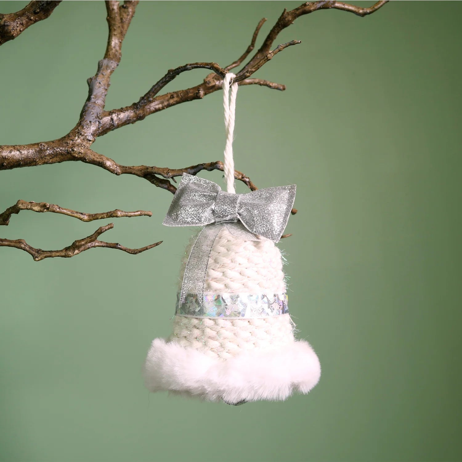 A white knitted Christmas bell ornament with a fluffy white faux-fur trim and a glittery silver bow, hanging from a brown tree branch against a solid sage green background.