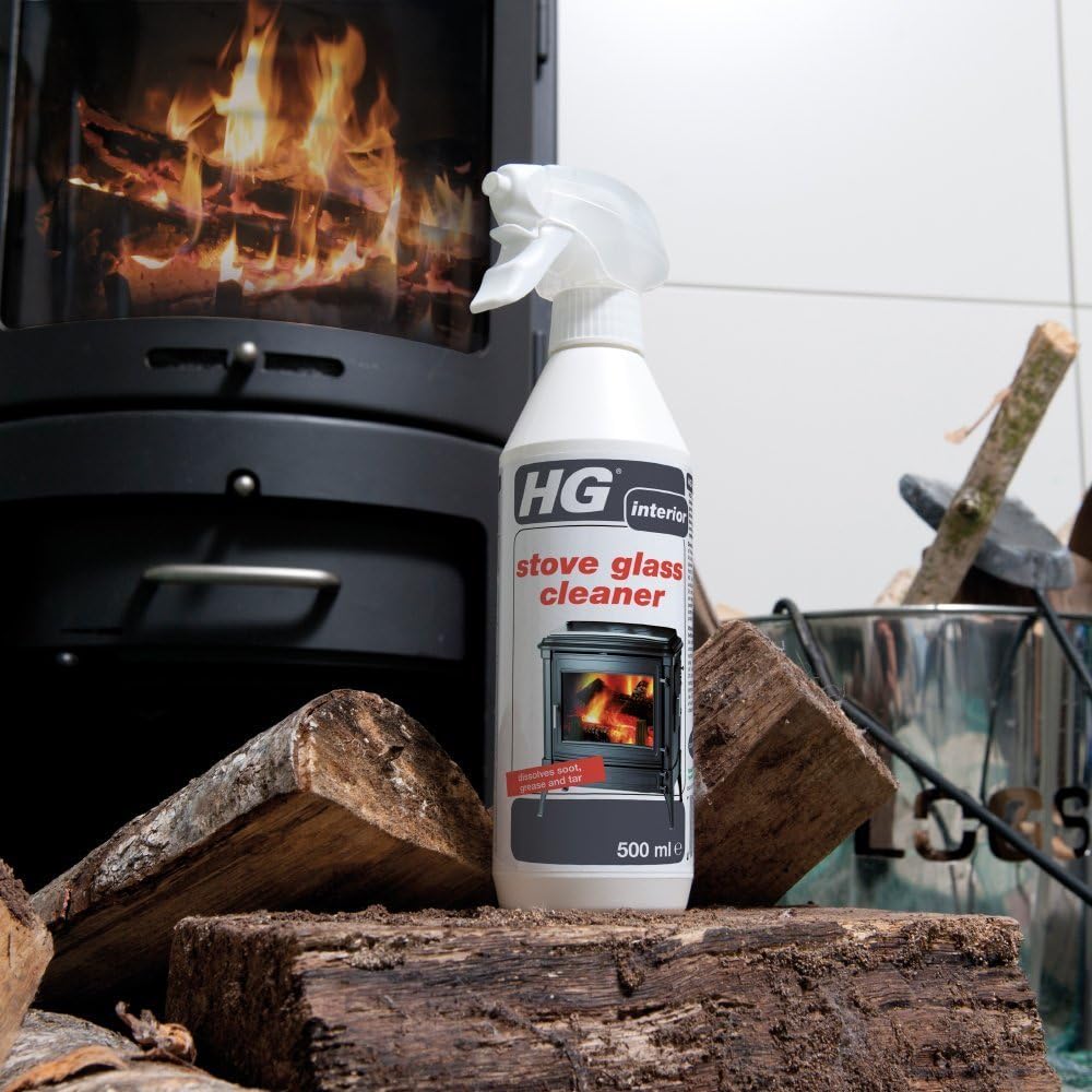 A bottle of HG Interior stove glass cleaner stands on stacked firewood in front of a black wood-burning stove with a visible fire, with a metal bucket and additional logs in the background.