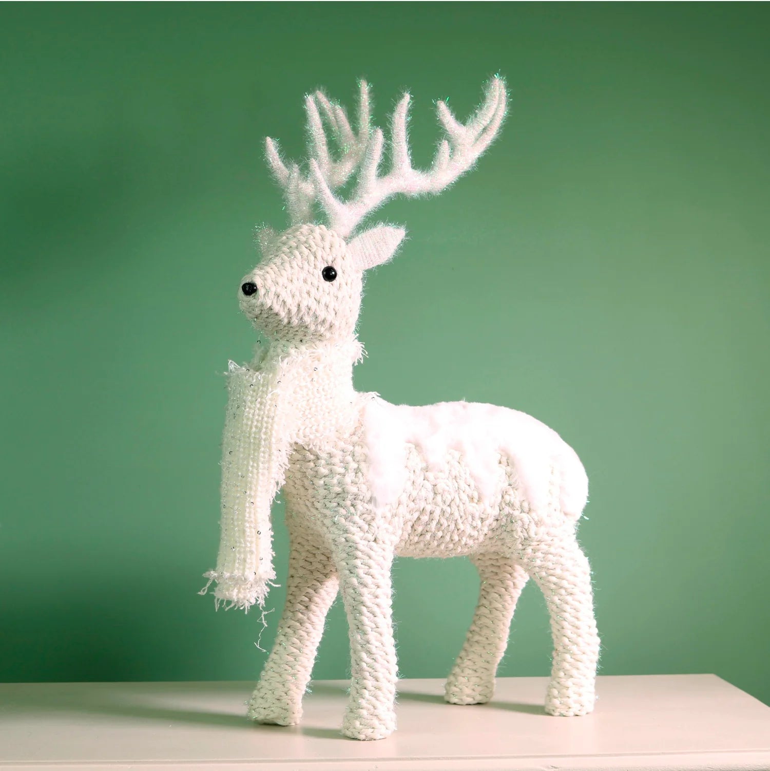 A white knitted reindeer figurine with antlers, standing on a light surface against a solid green background.