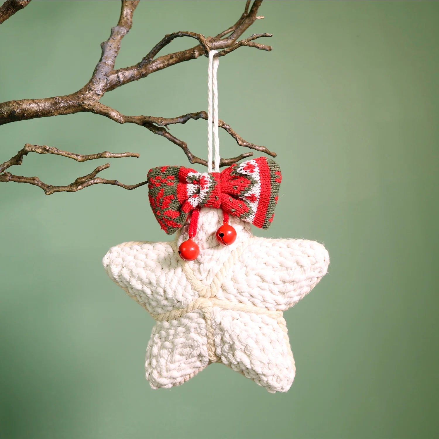 A crocheted star-shaped Christmas ornament in cream yarn, decorated with a red and green patterned bow, two red beads, and a white hanging string, shown hanging from a tree branch against a soft green background.
