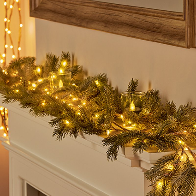 A festive pine garland with warm white string lights draped across a white fireplace mantel, decorated with small glass tealight candles, set against a neutral wall with a wooden mirror frame.
