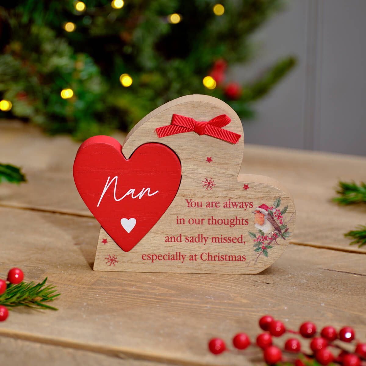A wooden Christmas ornament plaque shaped like two hearts, one red and one natural wood, displayed on a wooden table with a blurred Christmas tree in the background. The red heart is labeled 'Nan' and the wood heart has a red ribbon bow and printed holiday message.
