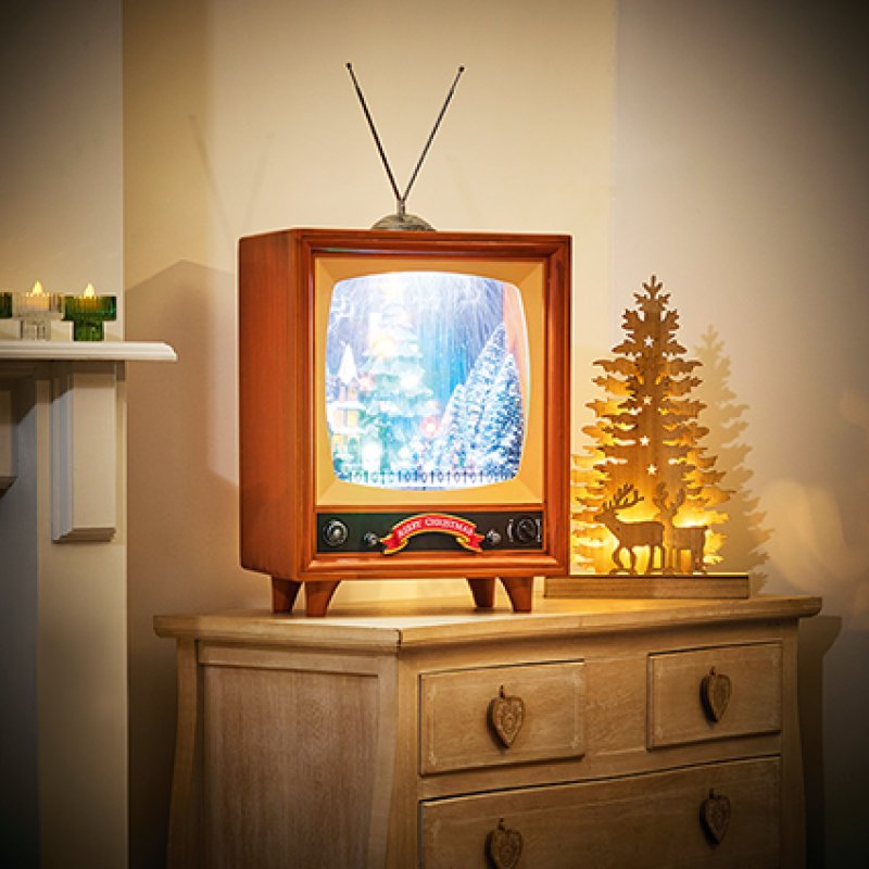 A retro-style wooden television lamp with a glowing winter forest scene on the screen, placed on a wooden dresser in a warmly lit room. A decorative tree silhouette with a deer sits beside it.
