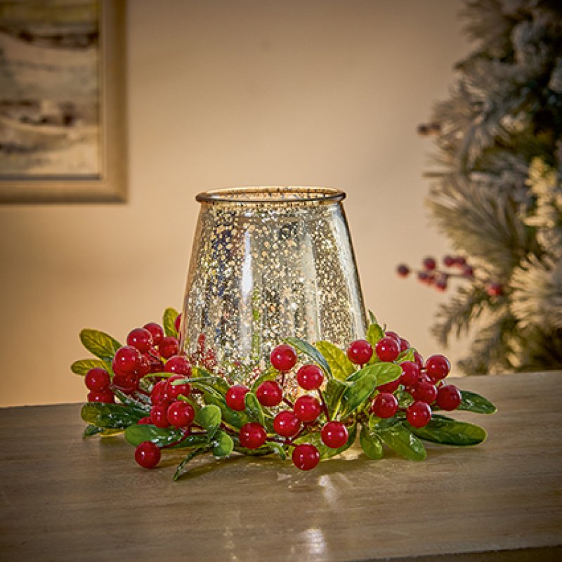 A decorative holiday centerpiece featuring a glittery silver inverted glass candle holder surrounded by a faux cranberry garland with red berries and green leaves, set on a wooden table with a softly blurred Christmas tree in the background.